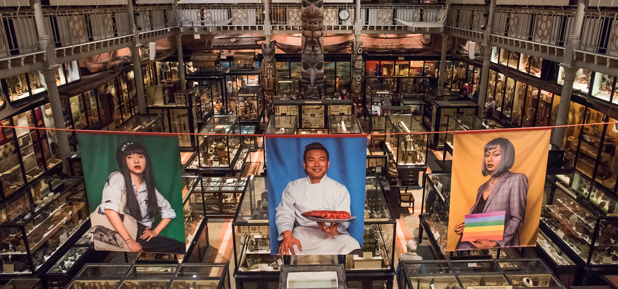 the central hall of the Pitt Rivers Museum with large flags showing colourful contemprary portraits of people