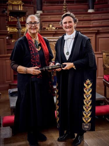 June Northcroft Grant and Irene Tracey standing in front of rows of wood-panelled seating, both holding a degree certificate in a ceremonial holder. Credit: Fisher Studios