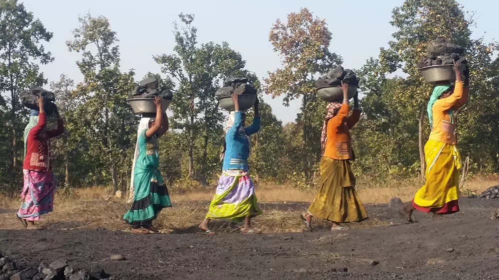 5 women carrying baskets of dark rocks on their heads