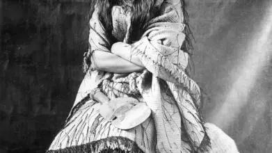 A black and white photo of Mākereti Papakura. She is sitting down with her arms folded, wearing a garment of loose, light-coloured fabric over a dark skirt.