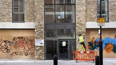 A worker in high vis completes work on a modern apartment building with boarded up graffitied shops