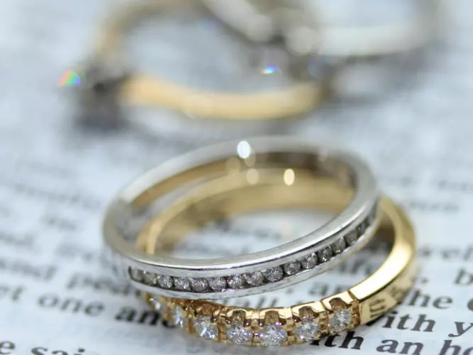 A silver diamond ring stacked on top of a gold diamond ring, resting on a page. In the background are two other rings.