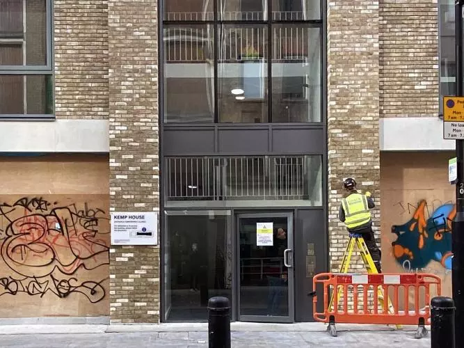 A worker in high vis completes work on a modern apartment building with boarded up graffitied shops