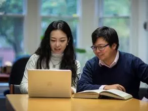 Two students sat together at a desk with a book and a laptop.