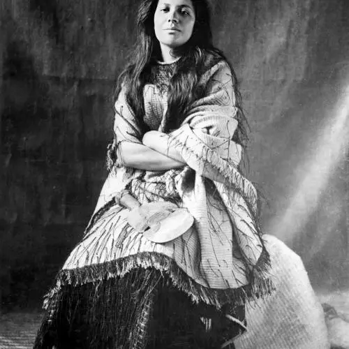 A black and white photo of Mākereti Papakura. She is sitting down with her arms folded, wearing a garment of loose, light-coloured fabric over a dark skirt.