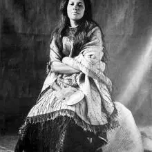 A black and white photo of Mākereti Papakura. She is sitting down with her arms folded, wearing a garment of loose, light-coloured fabric over a dark skirt.