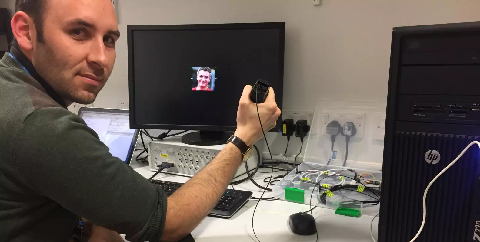 A man squeezes a black object whilst sitting at a desk with a small photo of another man displayed on the screeen
