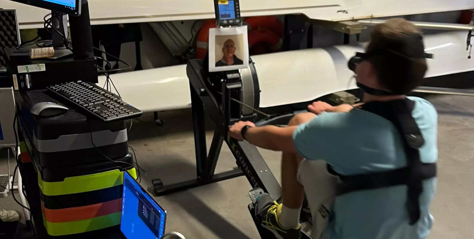 A participant rowing while viewing the photo of a support figure in a room filled with white rowing boats