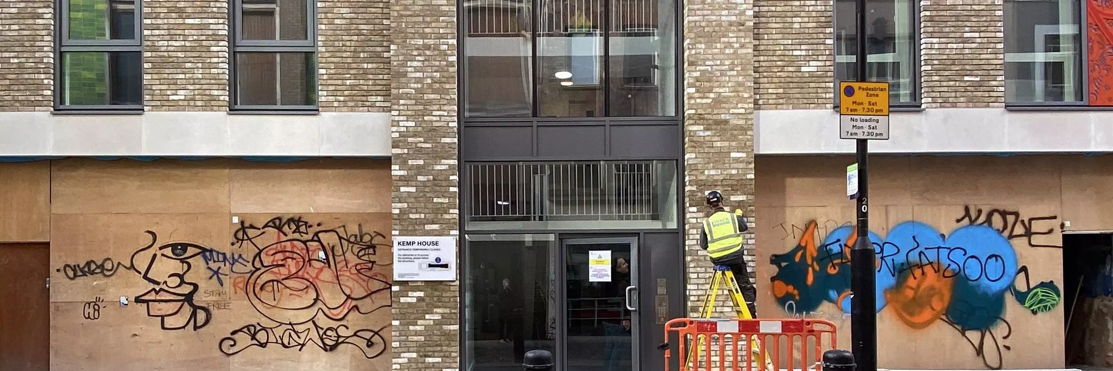 A worker in high vis completes work on a modern apartment building with boarded up graffitied shops