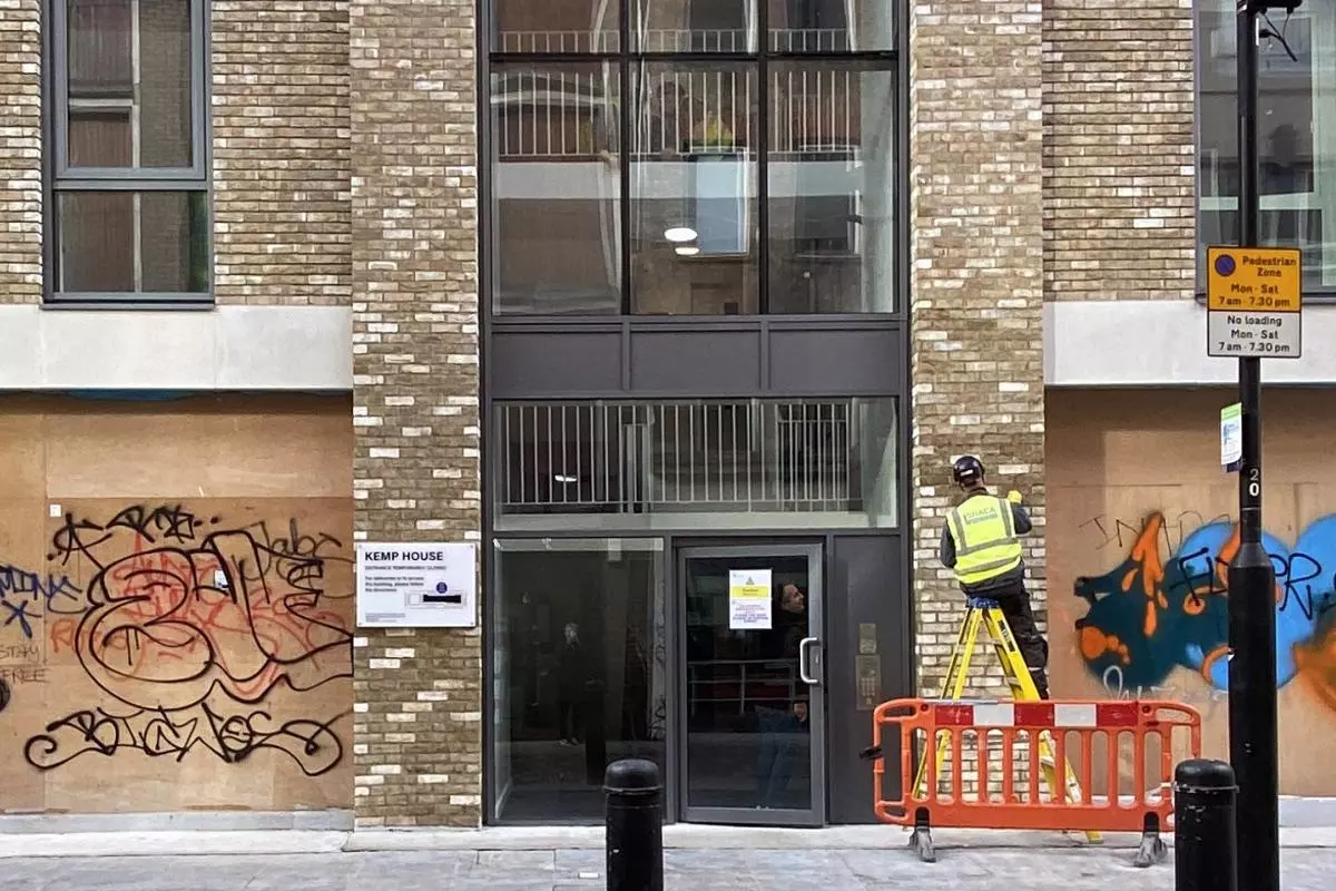 A worker in high vis completes work on a modern apartment building with boarded up graffitied shops